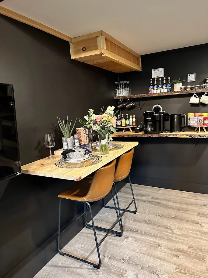Corner of a modern kitchen with black walls, a light wood breakfast bar set for two, a small floral arrangement, potted plants, wine glasses, and bar stools with tan seats. The backsplash has open shelves with glassware, bottles, coffee makers, and m