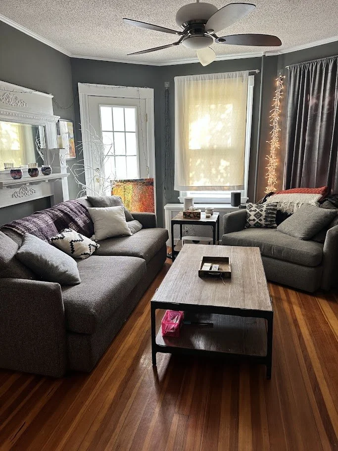 Living room with two gray couches, a wooden coffee table, white and dark curtains, and sunlight coming through a window and door.