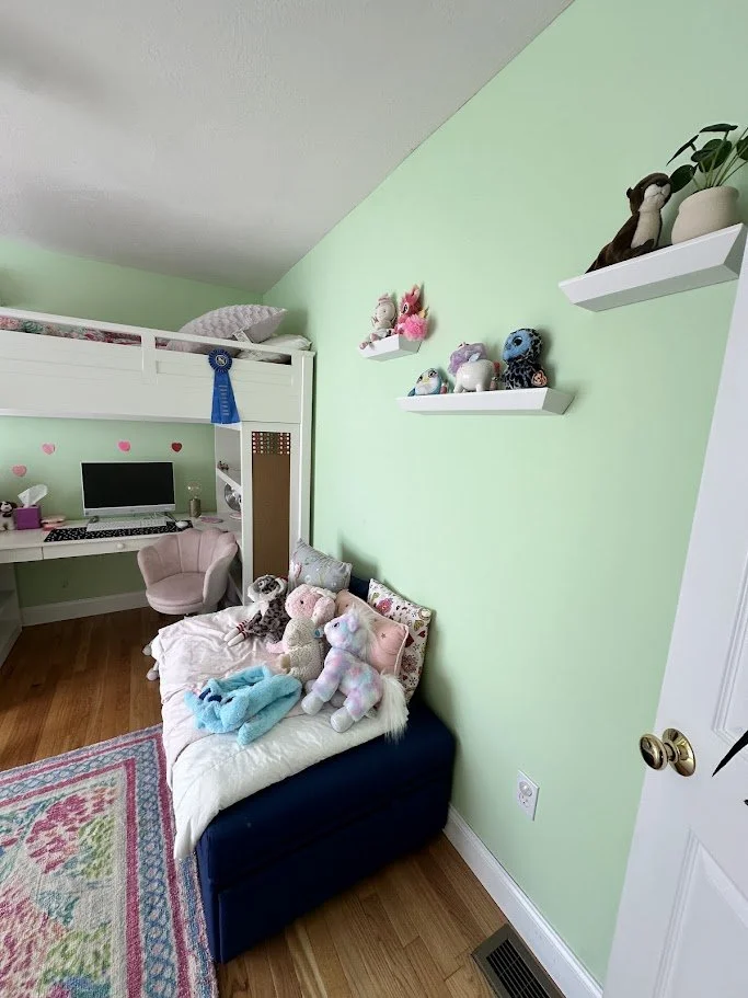 A cozy bedroom with a high bunk bed, a computer desk, and a pink armchair. A black storage bench holds stuffed animals, and plush toys are on the wall shelves. The room has mint green walls and wooden flooring.