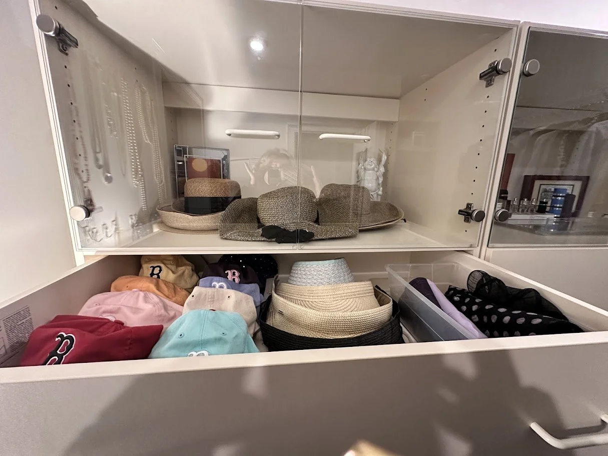 A display cabinet containing hats, caps, and accessories on two shelves. The top shelf has various hats in a glass cabinet, and the lower shelf has folded caps, hats, and miscellaneous items.