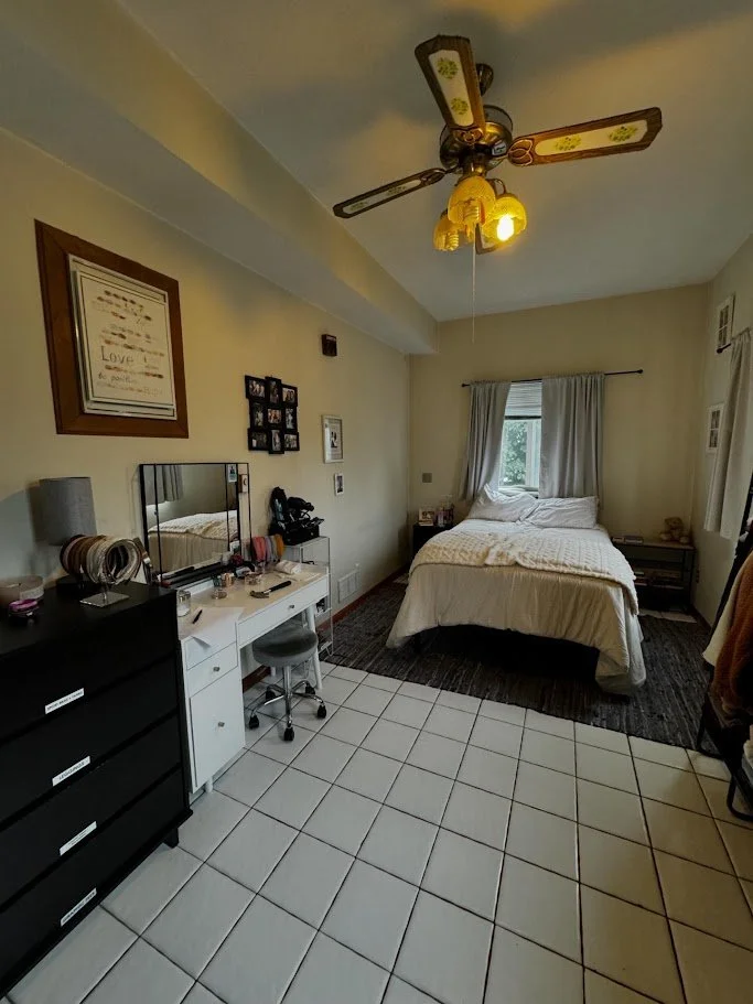 A bedroom with a bed, a window with curtains, a ceiling fan, a dresser, a mirror, and a small desk with a stool.