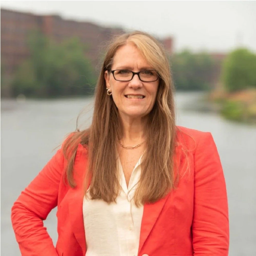 Woman with long hair and glasses wearing a red blazer, standing outdoors near a body of water.