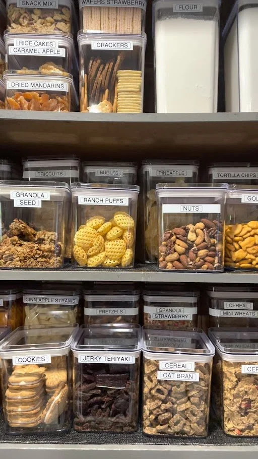 Shelf with various snack containers labeled with items such as rice cakes, caramel apple, crackers, flour, dried mandarins, granola seed, bugles, pineapple, tortilla chips, nuts, cinnamon apple straws, pretzels, cookies, jerky teriyaki, cereal, oat b