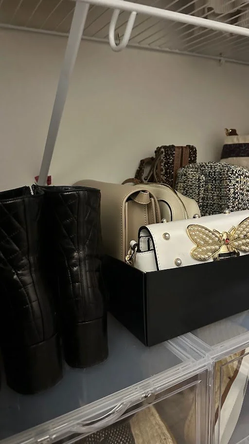 Black quilted boots and several handbags, including a cream purse with a butterfly embellishment, on a clear plastic container shelf.