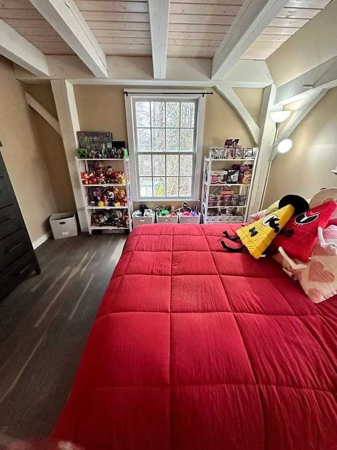 A cozy bedroom with a large bed covered with a red quilt, decorated with plush toys resembling cartoon characters. There is a window in the background flanked by two white bookshelves filled with toys and collectibles. The room has a wooden ceiling w