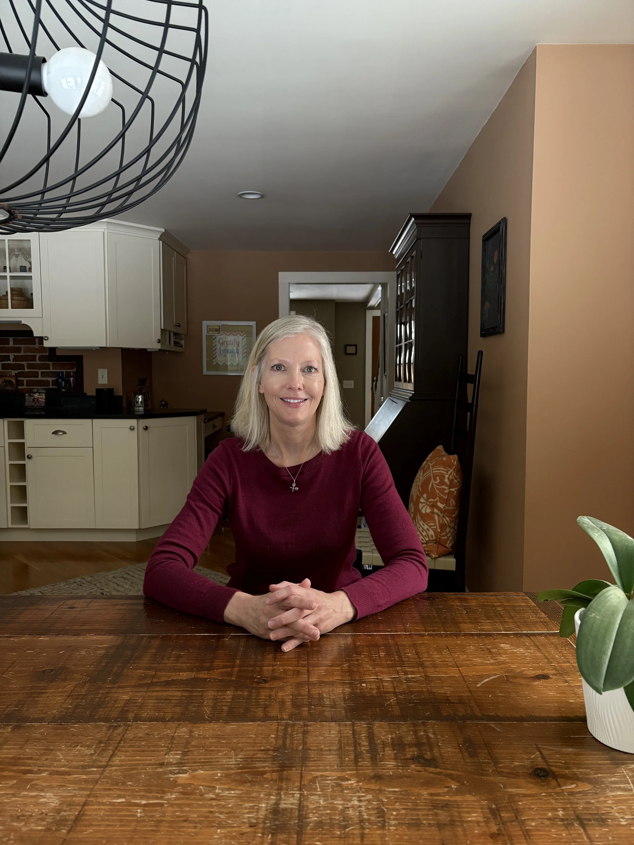 Woman with blonde hair sitting at a wooden table in a cozy kitchen with a plant in the foreground and a geometric light fixture above.