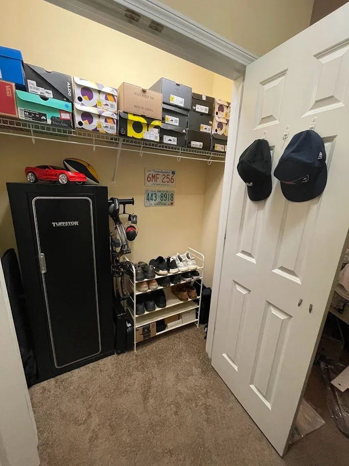 A small closet with a wire shelf holding shoe boxes, a shoe rack with various shoes, a gun safe, a small red toy car, two hats hanging on the door, and three license plates on the wall inside the closet.