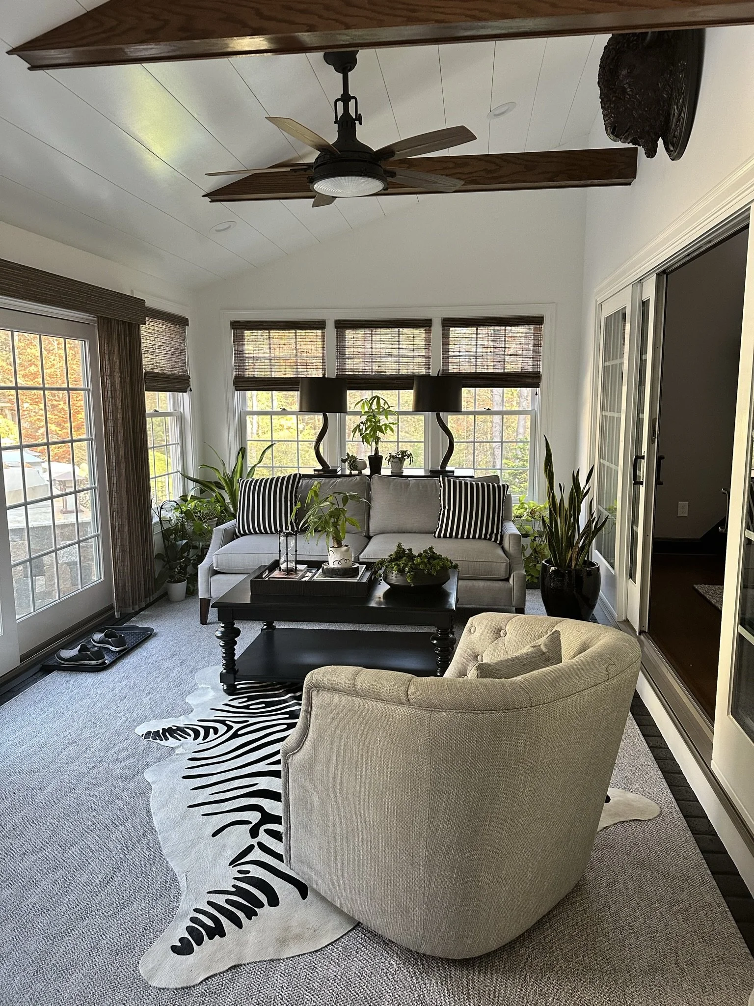 A sunroom with a cozy seating area featuring a gray sofa, a light-colored armchair, and a black coffee table. The room is decorated with plants, a zebra-patterned rug, and ceiling beams. There are large windows with bamboo shades, allowing natural light into the space.