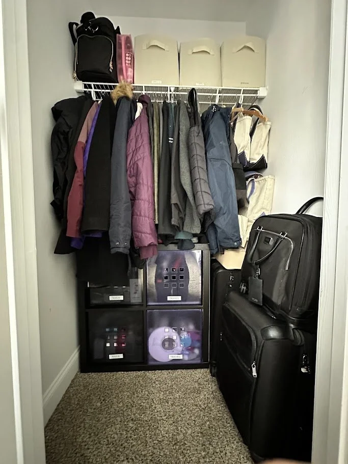 Small closet with hanging jackets and coats, plastic storage bins labeled 'Travel' and 'Shoes,' and suitcases on the floor.