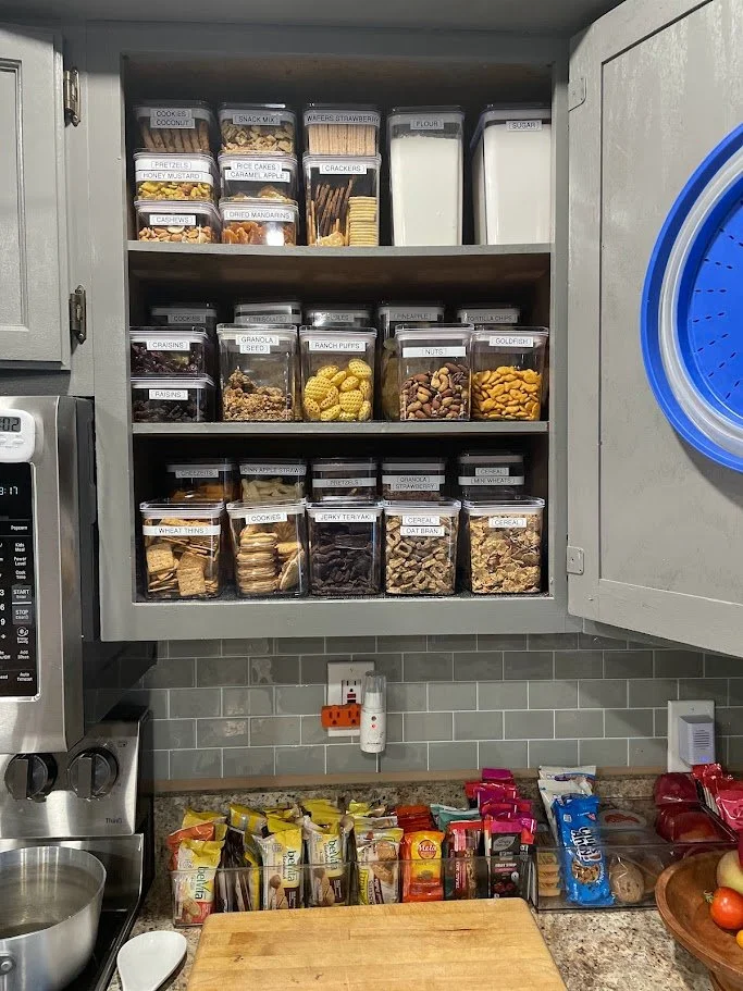 Top shelf contains snack jars labeled cookies, snack mix, wafers, and flour. Middle shelf has jars labeled craisins, raisins, granola, ranch puffs, and mixed nuts. Bottom shelf contains cookies, jerky, cereal, and bread. On the counter are various sn