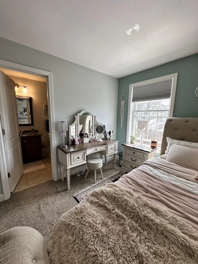 A cozy bedroom with a large bed, fluffy blanket, bedside table with a lamp, a vintage-style vanity with mirror, and a window with blinds looking outside. A small stool is in front of the vanity, and a doorway leads to a bathroom.