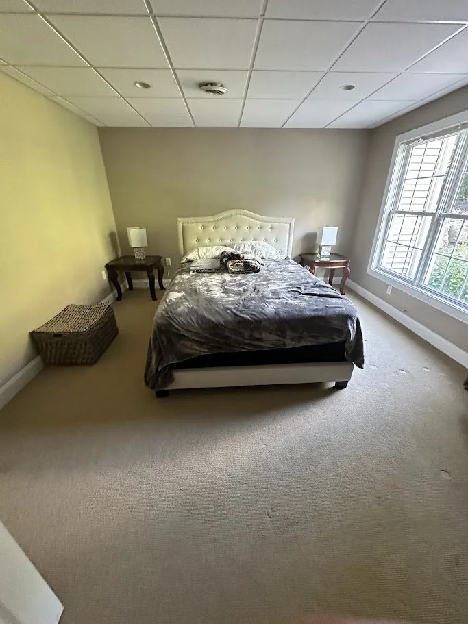 A bedroom with a bed, two nightstands with lamps, a wicker basket, and a large window.