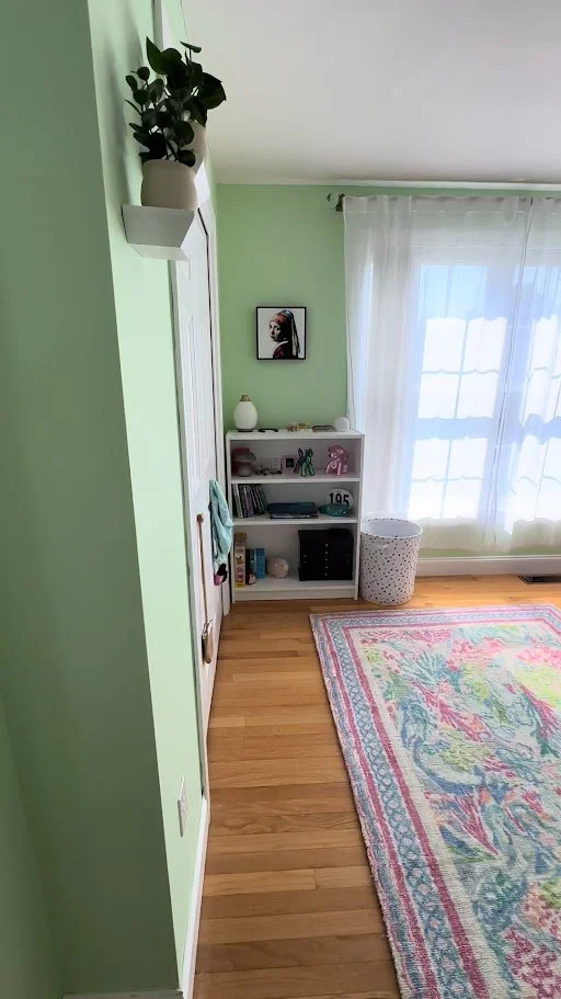 Corner of a room with a green wall, white shelf with books and decorations, a framed portrait on the wall, a window with white curtains, a white laundry basket with holes, and a colorful area rug on hardwood floor.