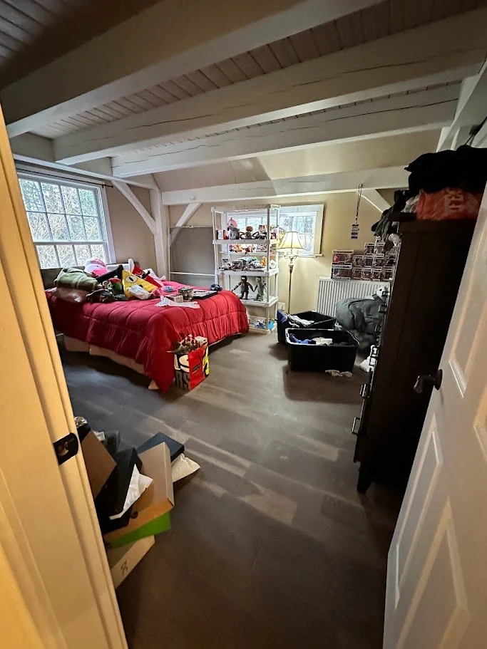 A bedroom with a red bedspread, toys on the bed, and various items stored on shelves and in boxes, illuminated by natural light from windows and a standing lamp.