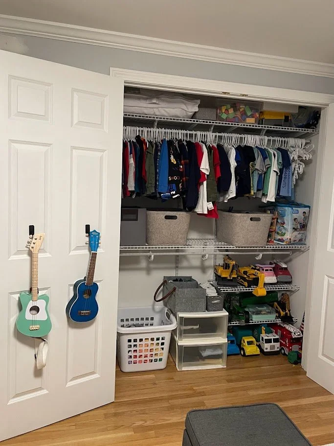 Open closet with organized children's clothes on a top rod, toy storage boxes, and a shelf with toy vehicles. Two small guitars hang on a door in front of the closet.