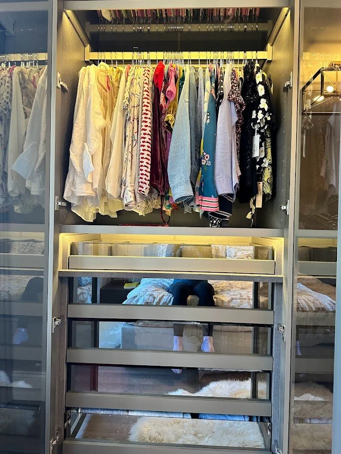 Open closet with hanging clothes and a mirror reflecting a person taking a selfie inside a bedroom.
