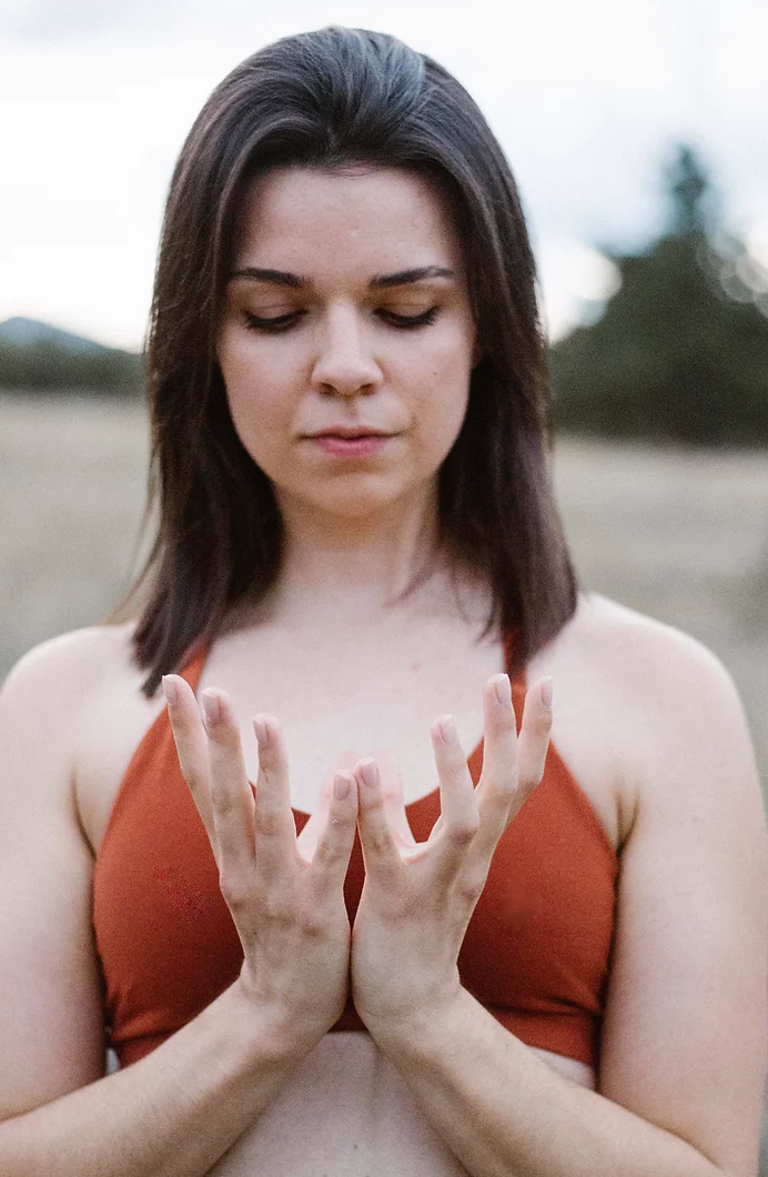 Woman in orange top with eyes closed, hands raised, serene outdoor setting.