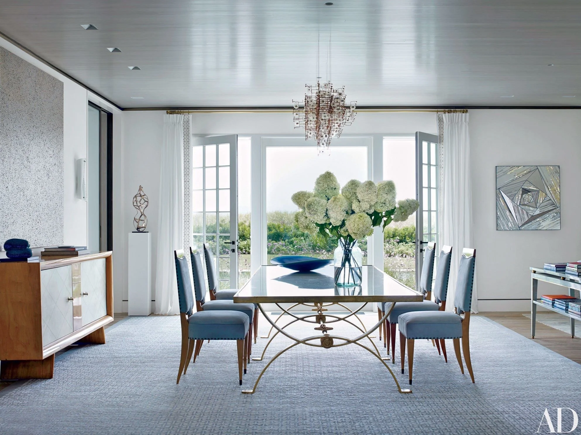 Elegant dining room with a large wooden table, blue upholstered chairs, a chandelier, white curtains, glass doors opening to a garden, and a large bouquet of white hydrangeas on the table.