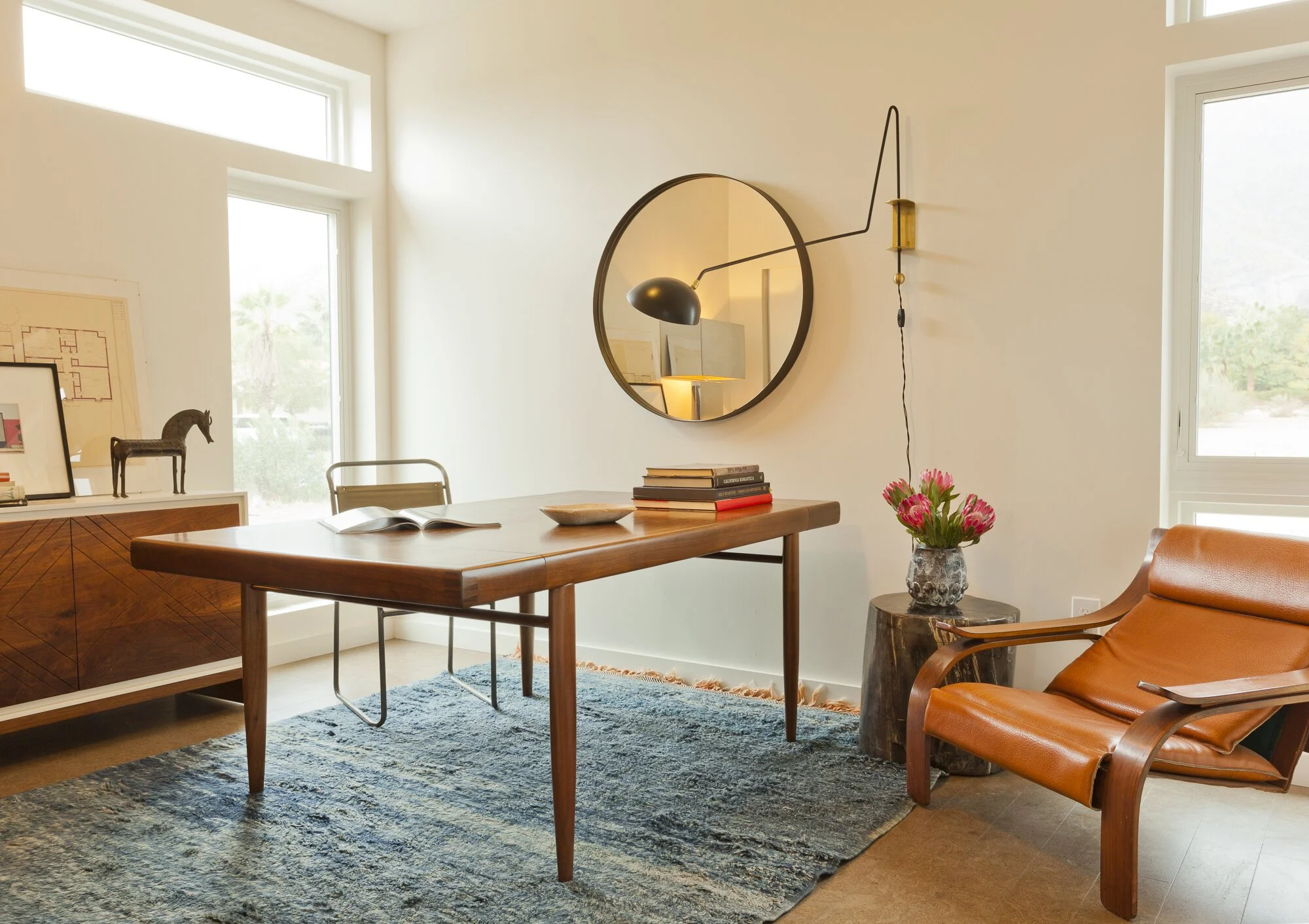 Modern living room with wooden dining table, leather lounge chair, round wall mirror, books, flower vase, window, and wall-mounted lamp.