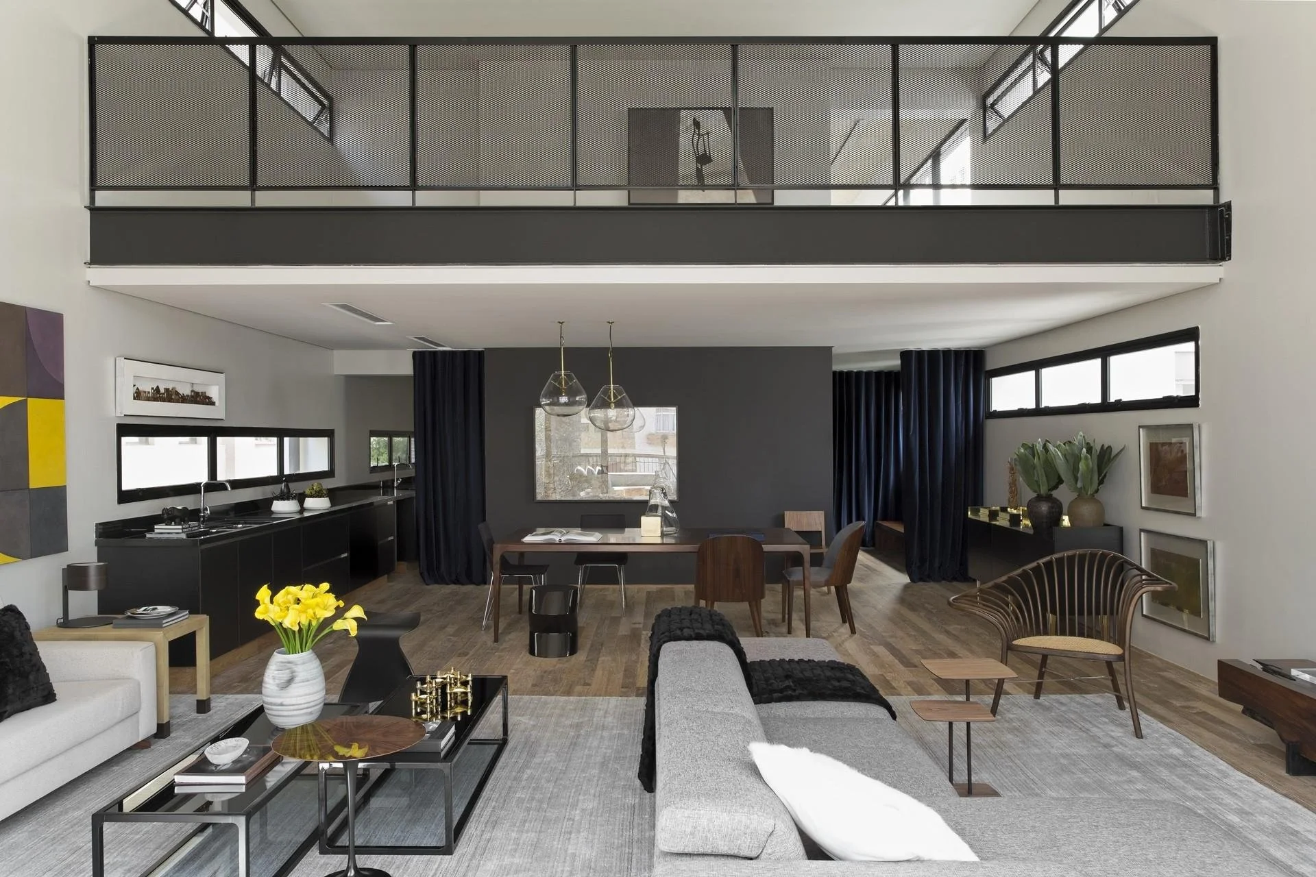 Modern living room with a grey sectional sofa, black coffee table, wooden dining table, black curtains, and an upstairs loft with metal railing.