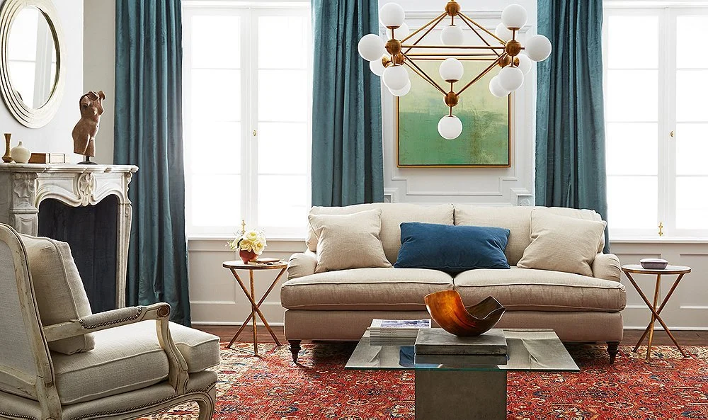 Living room with cream-colored sofa and armchair, blue and beige pillows, large windows with teal curtains, a modern chandelier, a fireplace mantle, a coffee table with a decorative bowl, and artwork on the wall.