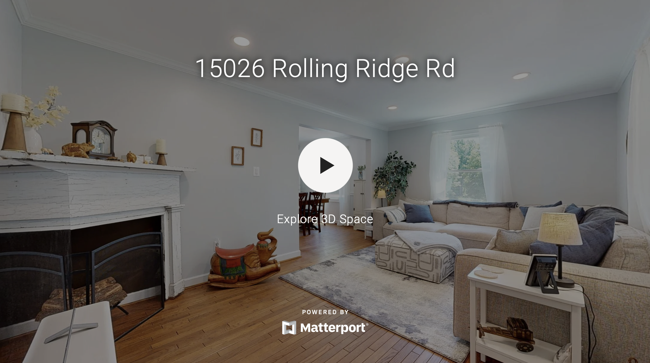 Living room with white walls, hardwood floors, a beige sectional sofa with blue and white pillows, a side table with a lamp and picture frame, a carpet, a fireplace with decorative items on mantel, and windows with white curtains.