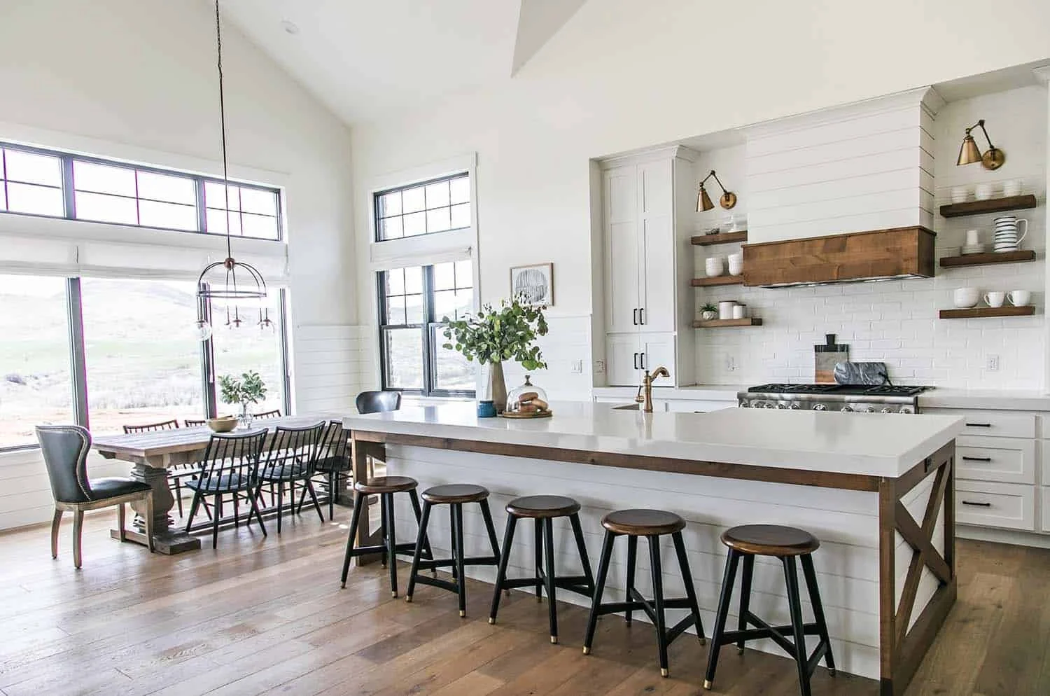 Modern kitchen with white cabinets, wooden accents, and large windows, featuring a central island with bar stools and a dining area with a wooden table and mixed chairs.