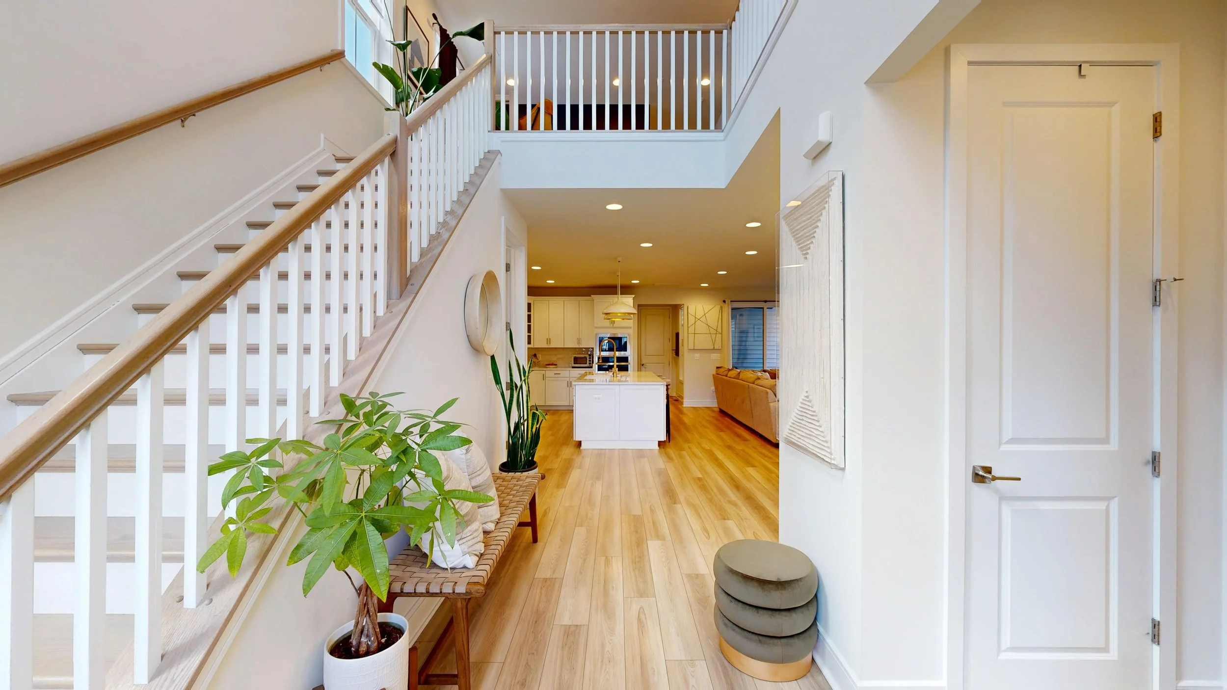Interior view of a modern home featuring a staircase with wooden handrails, a sitting area with a plant and a bench, a glimpse of a kitchen with white cabinets and an island, and open space leading to a living room with a couch.