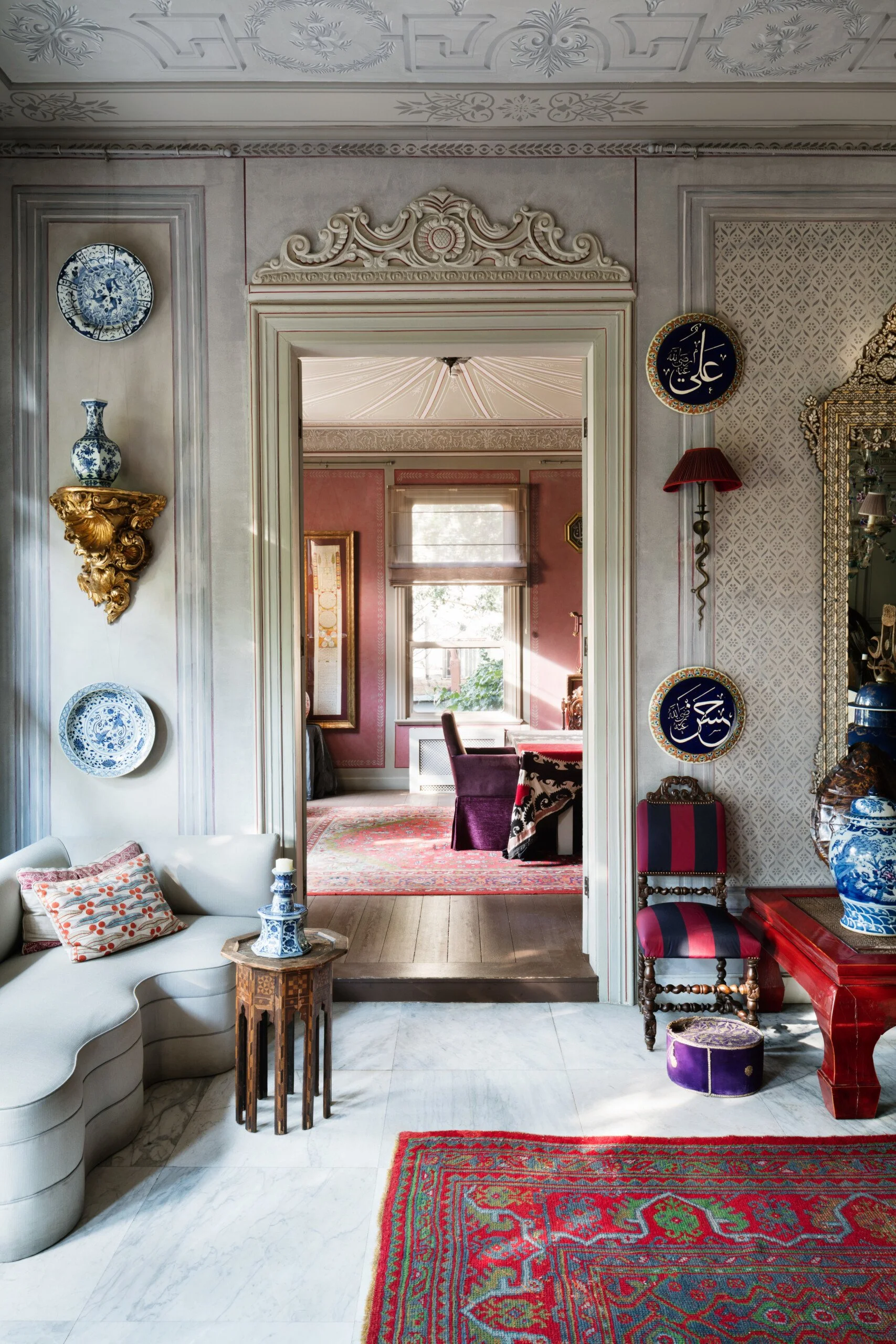 Elegant interior of a home with ornate architectural details, decorated with blue and white porcelain plates and vases, with a glimpse into a pink and purple furnished dining room through a doorway.