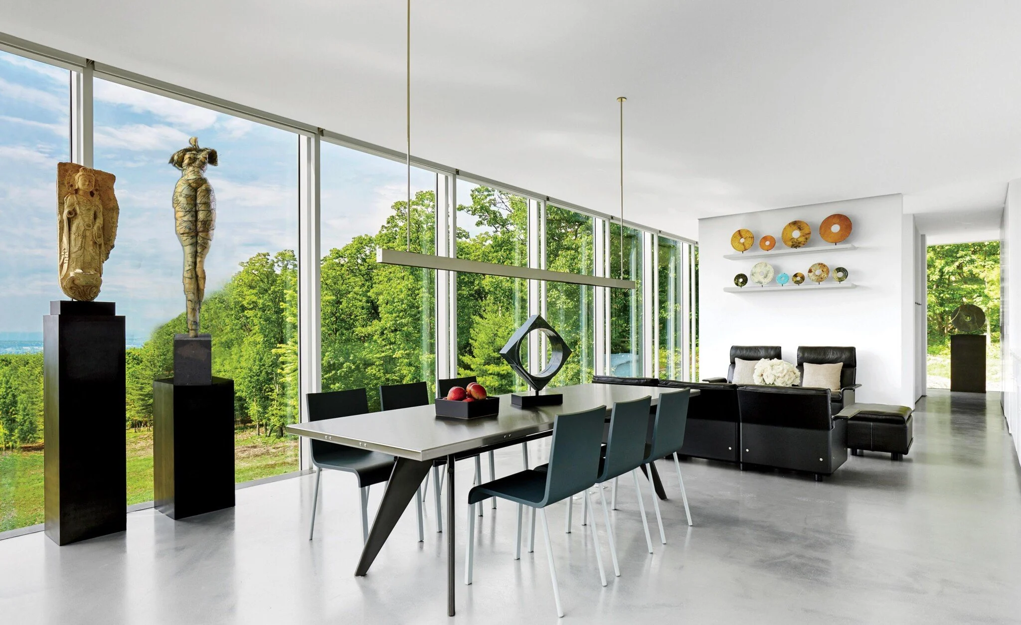 Modern living room and dining area with large floor-to-ceiling windows showing a lush green landscape, featuring sculptures and decorative wall art.