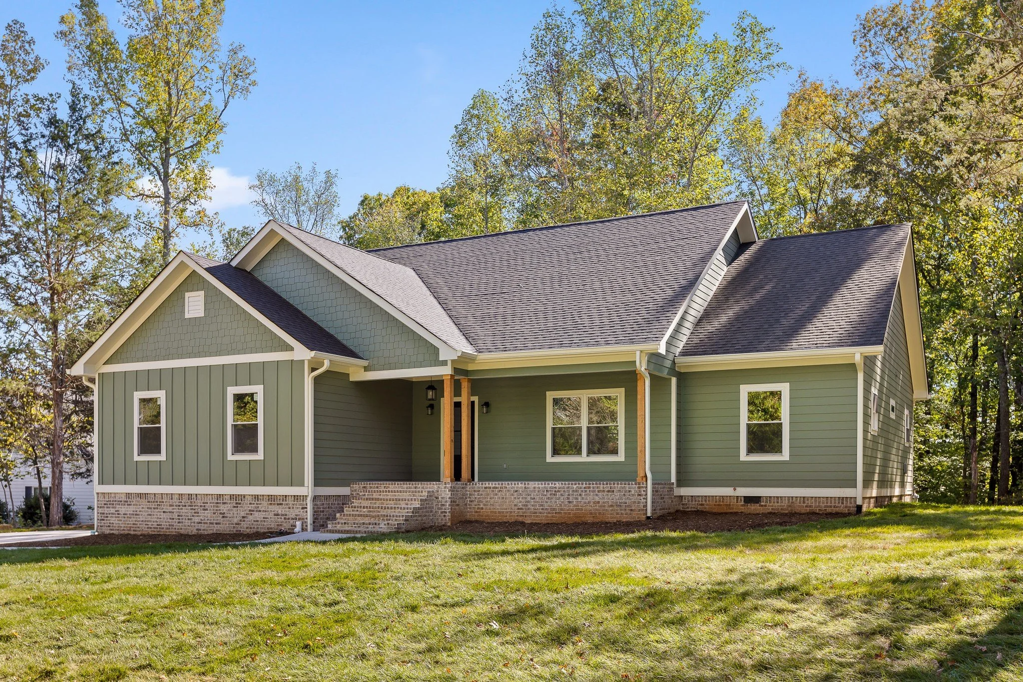 A newly constructed house with green siding, brick foundation, and dark gray roof, surrounded by trees and a grassy yard.