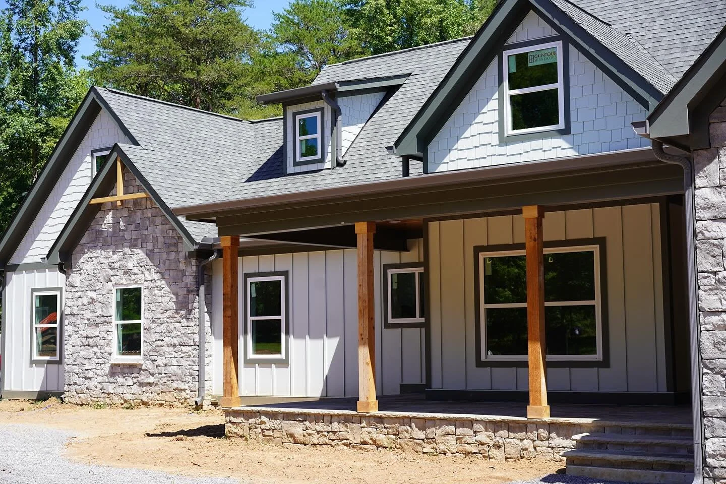 A house under construction with a porch supported by wooden beams, gray shingles, white siding, multiple windows, and a partly finished front yard.