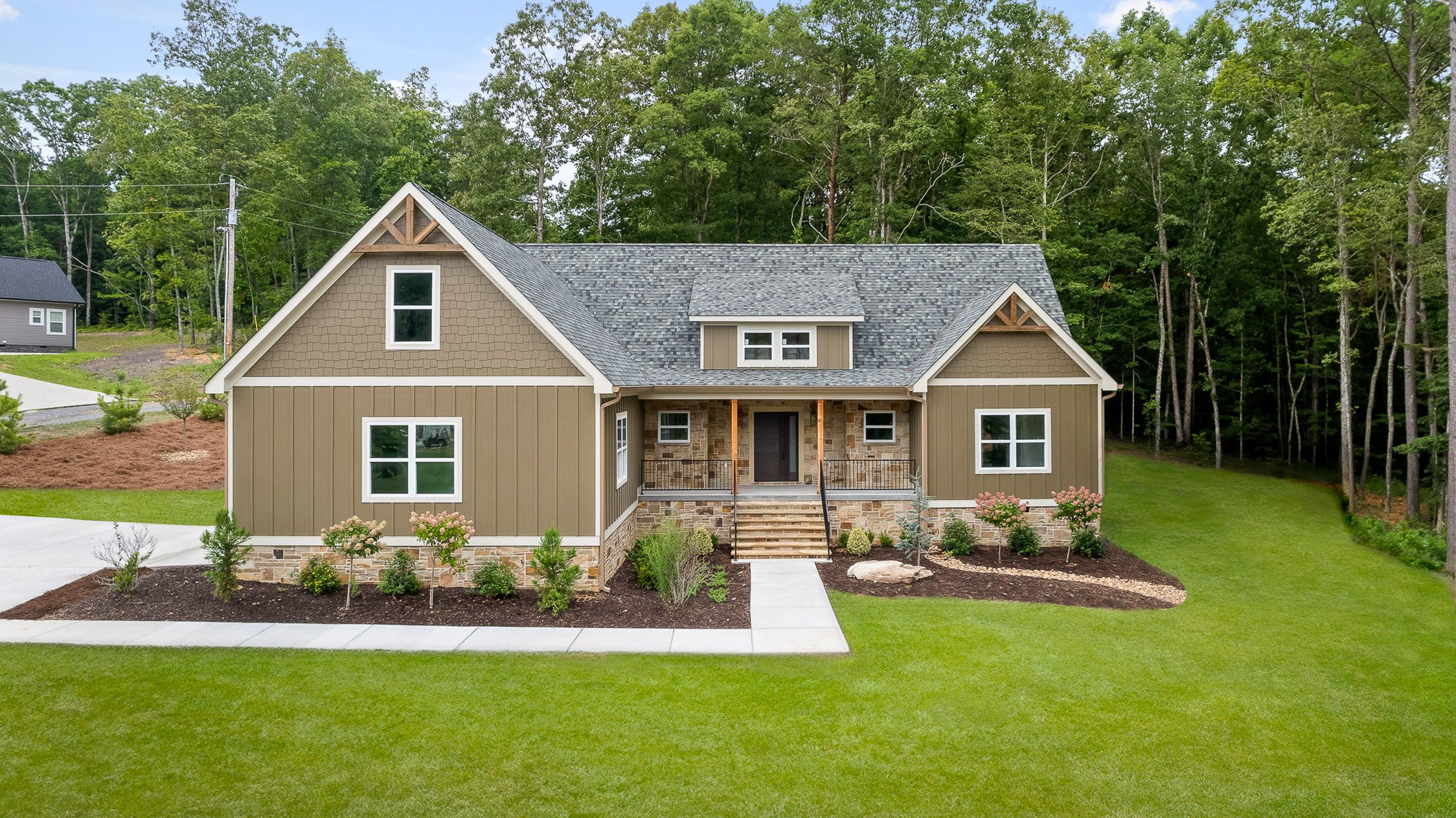 Front view of a newly built house with tan siding, stone accents, and a gray shingled roof, surrounded by well-maintained lawn and landscaped gardens, with a wooded area in the background.