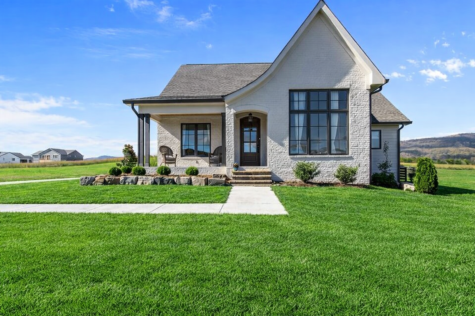 A white house with a steeply pitched roof, large front windows, and a small front porch with two chairs. The house is surrounded by a well-manicured green lawn with a sidewalk leading to the front steps. The background features a bright blue sky and distant hills.