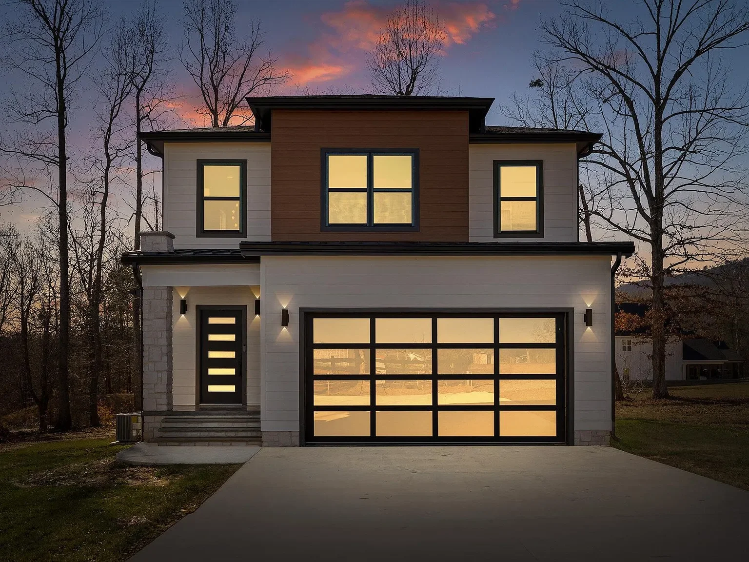 Modern two-story house with white and brown exterior, large glass garage door, front door with horizontal window slats, outdoor lighting, and bare trees in the background during sunset.