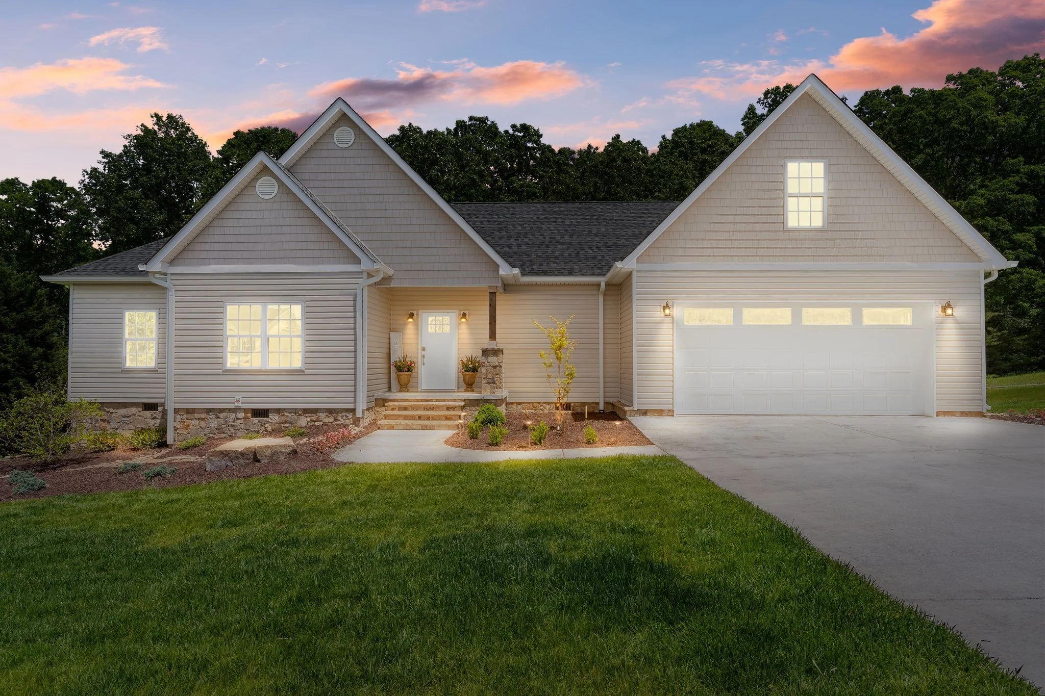 A modern suburban house at dusk with a well-manicured lawn, illuminated windows, and a two-car garage, surrounded by trees.