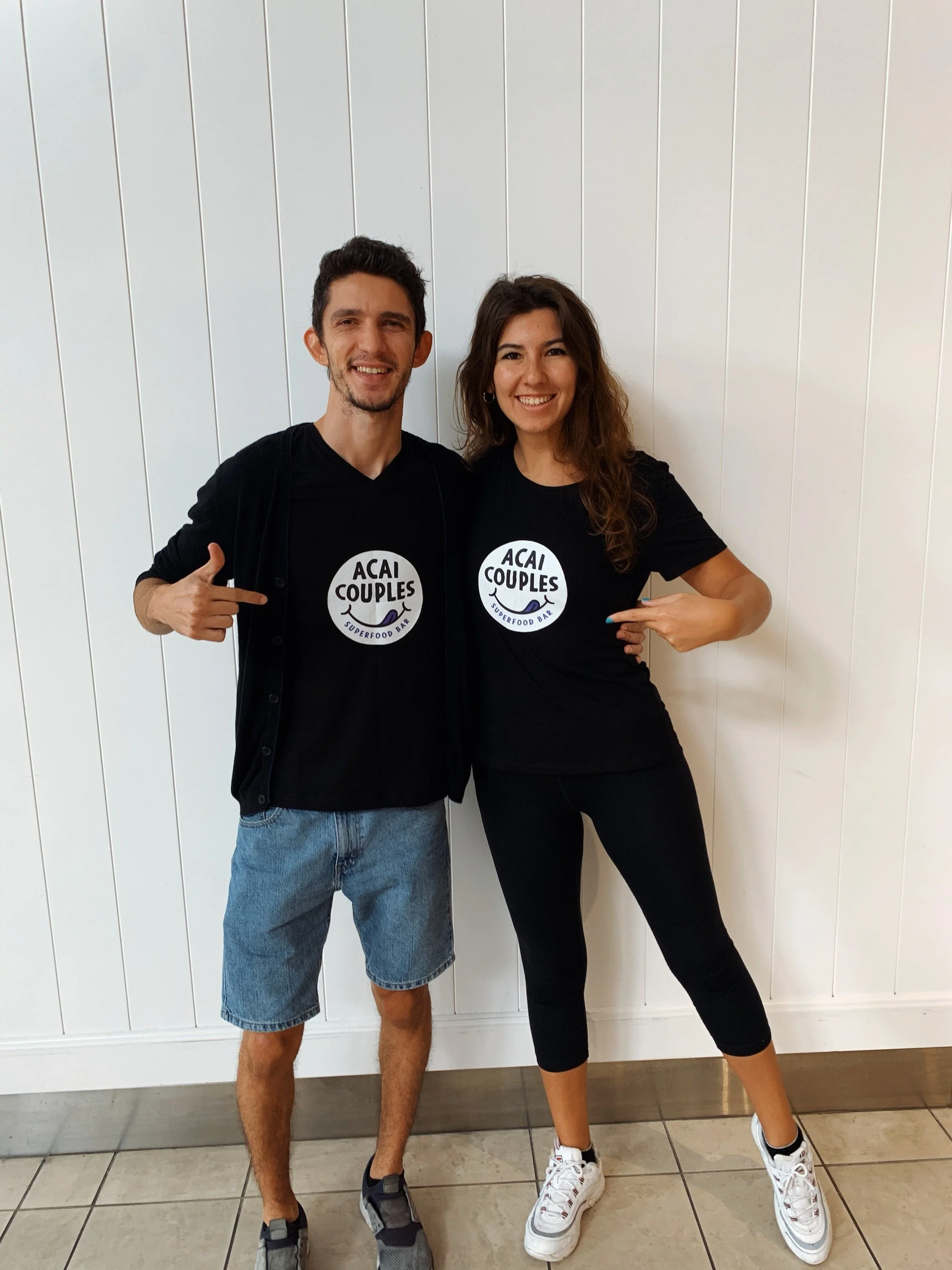 A happy young man and woman standing against a white wall, both pointing to their matching black t-shirts that say "ACAI COUPLES" with a small illustration of an acai bowl and the words "SUPERFOOD BAR" underneath in Myrtle Beach.