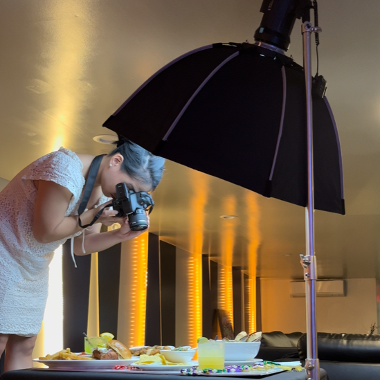 A woman taking a photograph of food on a table under a large black umbrella-shaped softbox light in a photography studio.