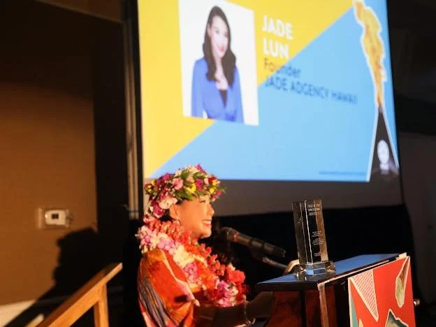 A woman wearing a floral lei and a flower crown speaking at a podium during an award ceremony. There is a large screen behind her displaying her photo and text related to the event.