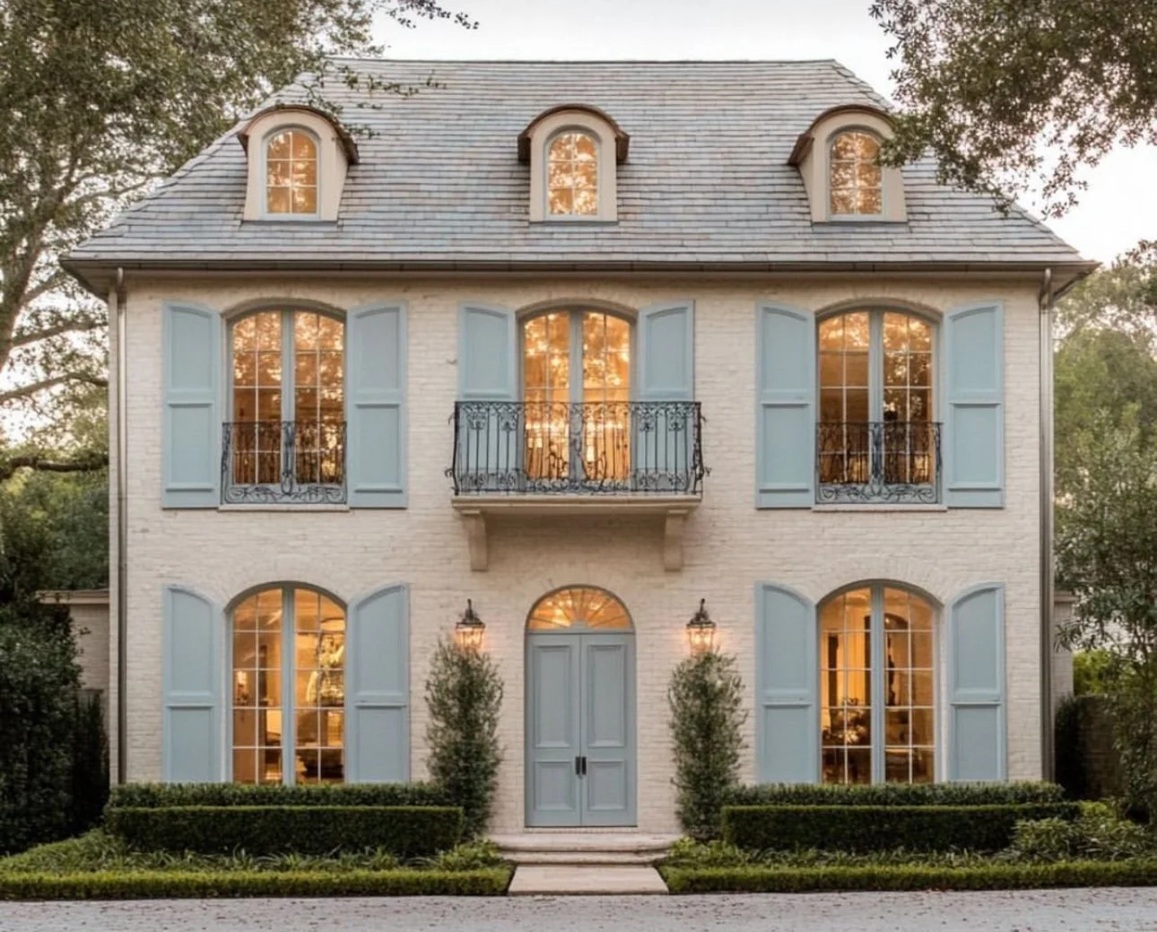 A three-story house with a white brick exterior, blue shutters, arched windows, and small balconies. The front has two lantern-style lights, two bushes, and a manicured hedge.