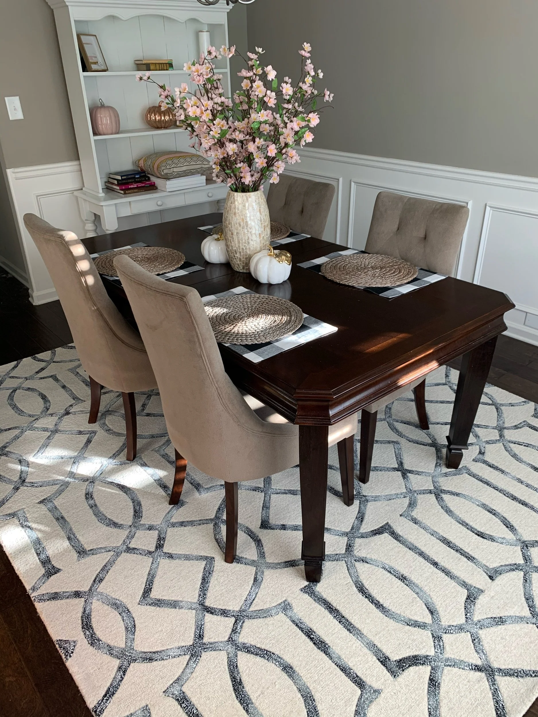 Dining room with a dark wood table, beige chairs, woven placemats, and a vase of pink flowers. White decorative pumpkins adorn the table. A white hutch is in the background, and the floor has a patterned rug.