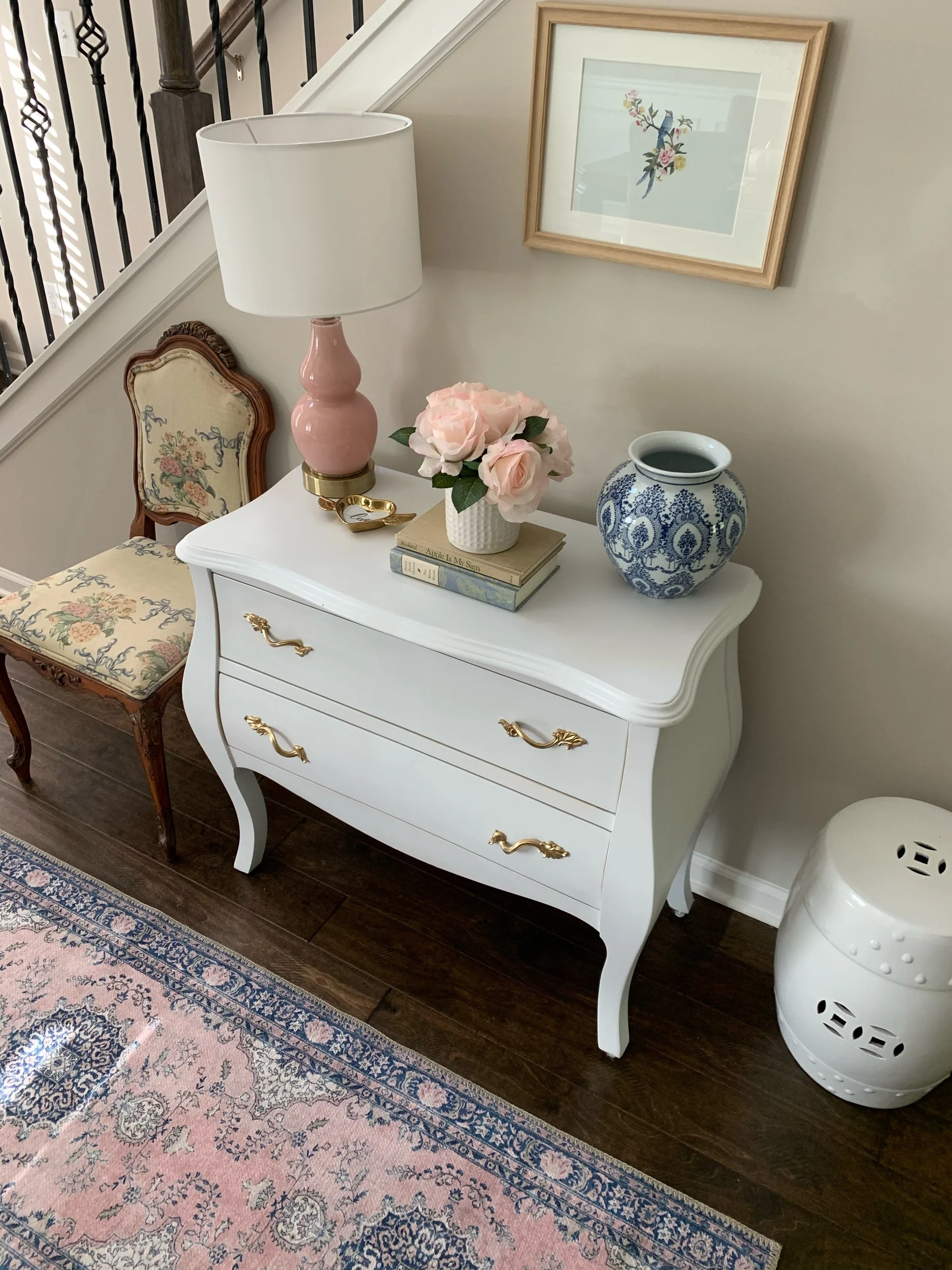 White dresser with gold handles, pink lamp, vase with pink roses, blue and white ceramic vase, framed floral artwork, floral-patterned chair, and a pink decorative rug on dark wood floor.