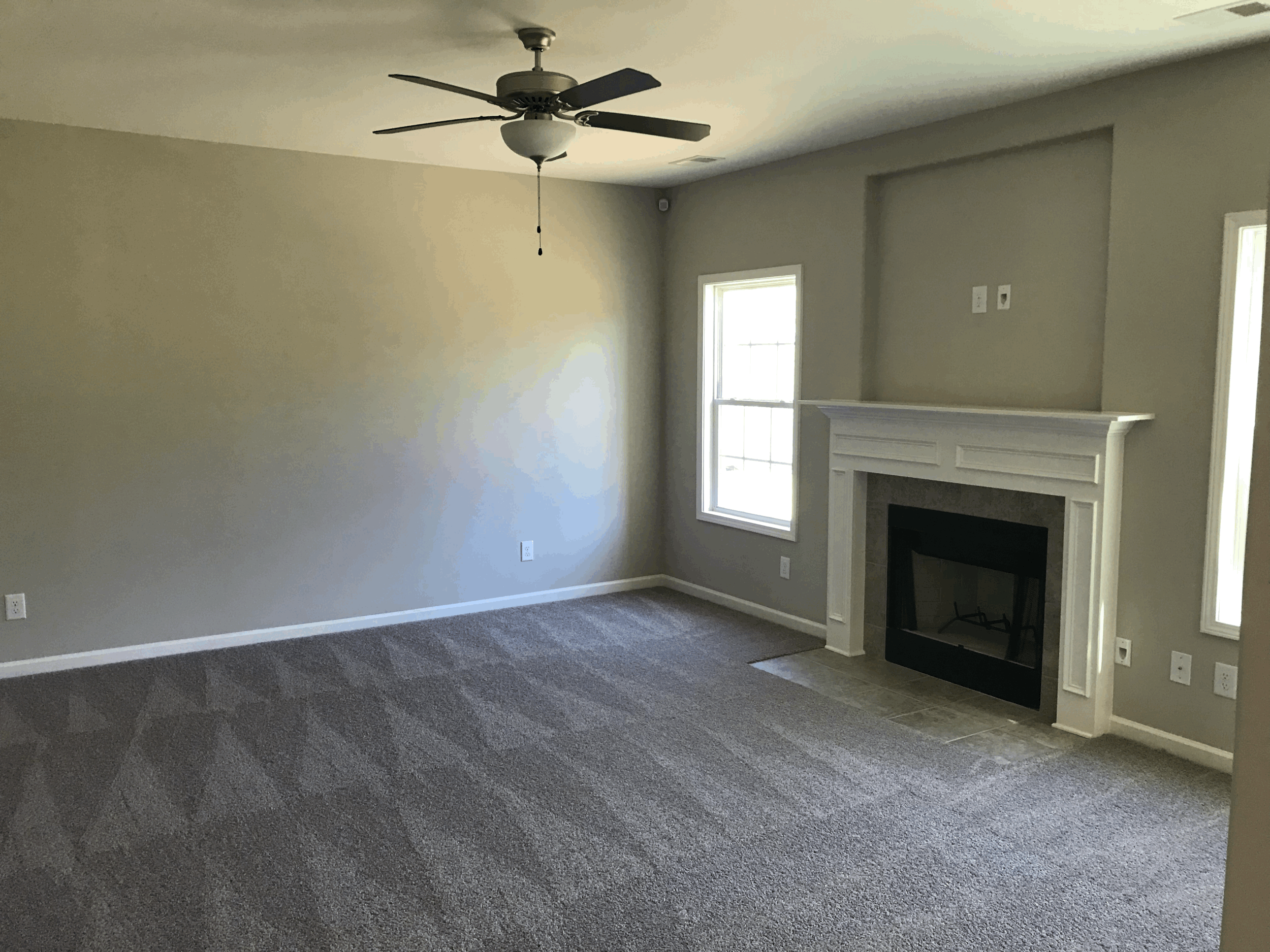 Empty living room with carpeted floor, ceiling fan, and a fireplace with white mantel next to two windows.