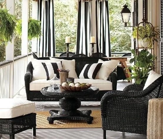 Elegant outdoor patio with black wicker furniture, white cushions, striped curtains, and potted plants. The decor features a table with a centerpiece and candles, placed on a woven rug.