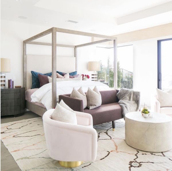 Modern bedroom with a canopy bed, pink and purple pillows, light pink armchair, small sofa, round coffee table, and abstract patterned rug. Large windows provide natural light.