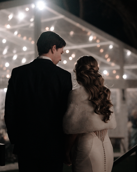 A couple at a nighttime event, standing close under string lights, facing away from the camera, with the man in a dark suit and the woman in a light-colored dress with long, curly hair.