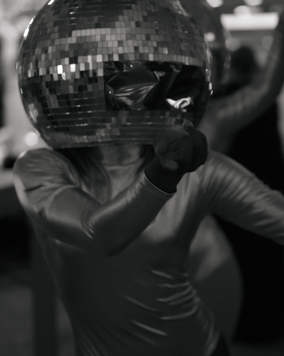 Person wearing a shiny, reflective helmet or headgear that resembles a disco ball, with a dark, long-sleeved shirt, indoors with blurred background.
