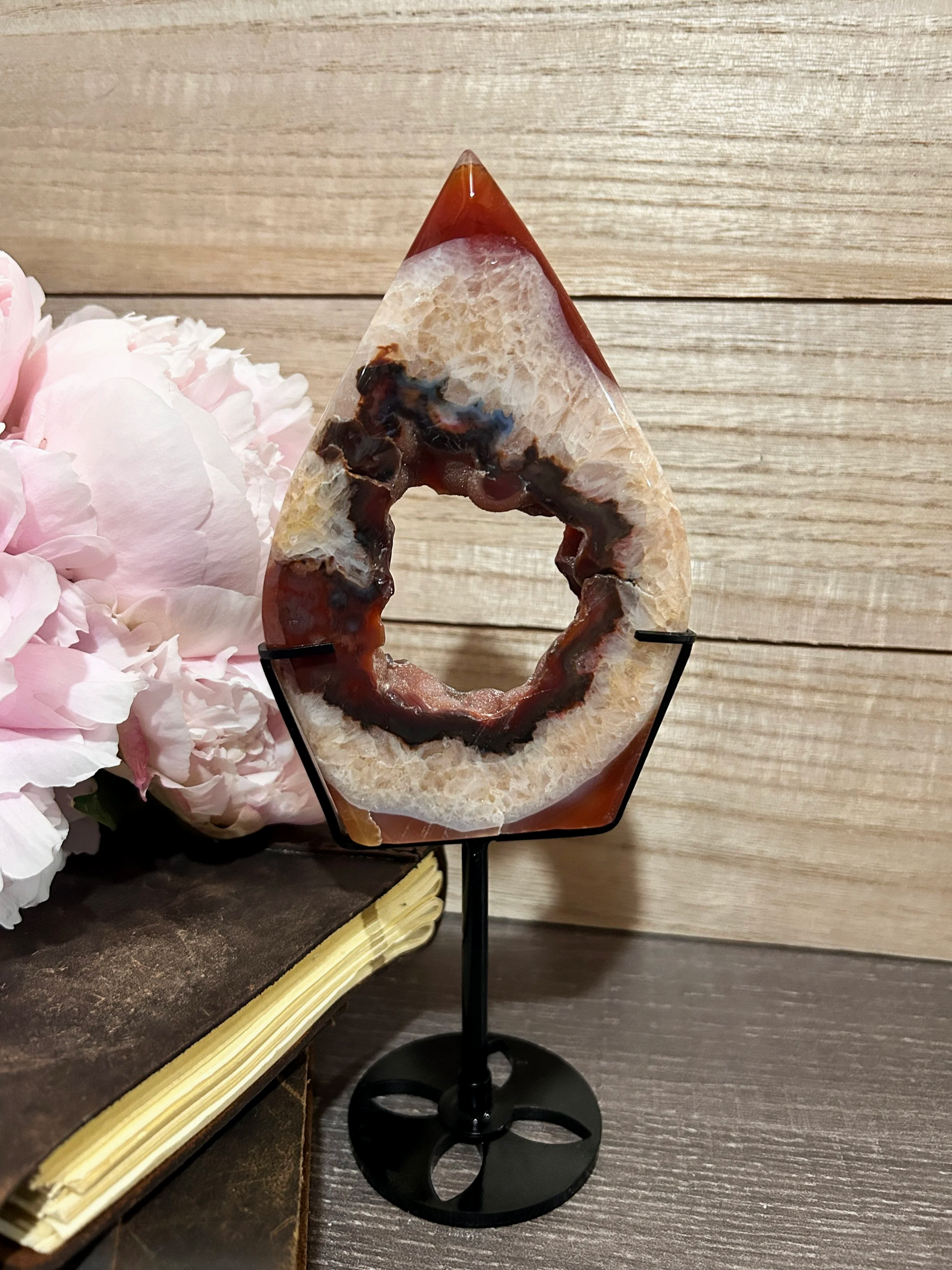 Decorative piece made of polished stone and resin on a black metal stand, placed on a wooden surface with pink flowers beside it.