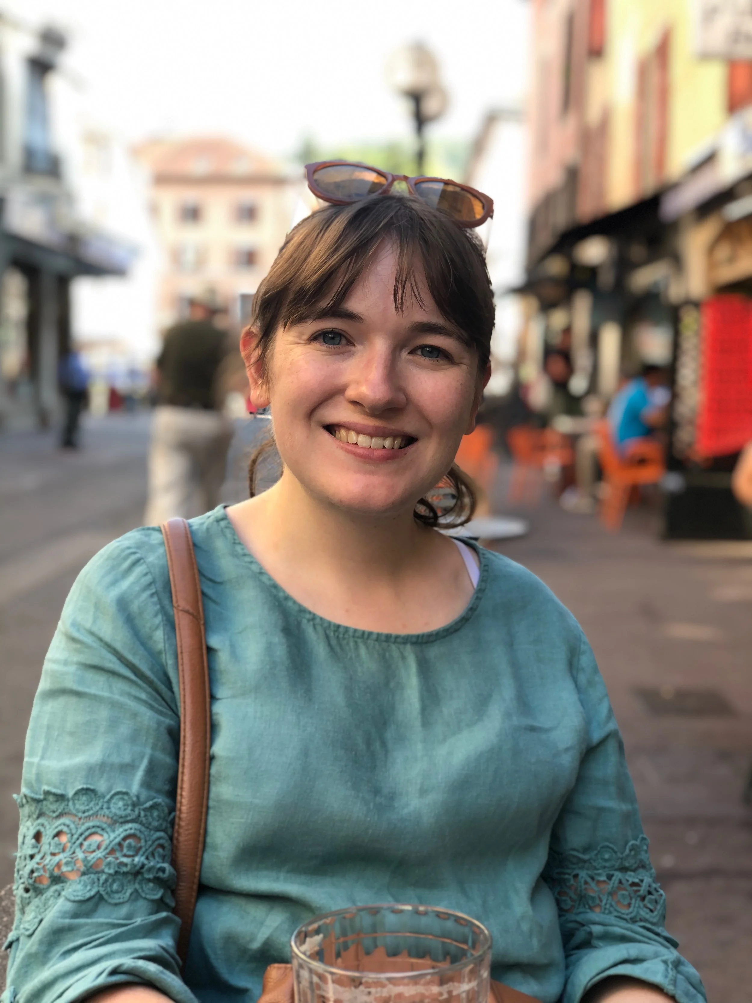 Person sitting at an outdoor cafe table, smiling, wearing a blue top and sunglasses on head, with a blurred street scene in the background.