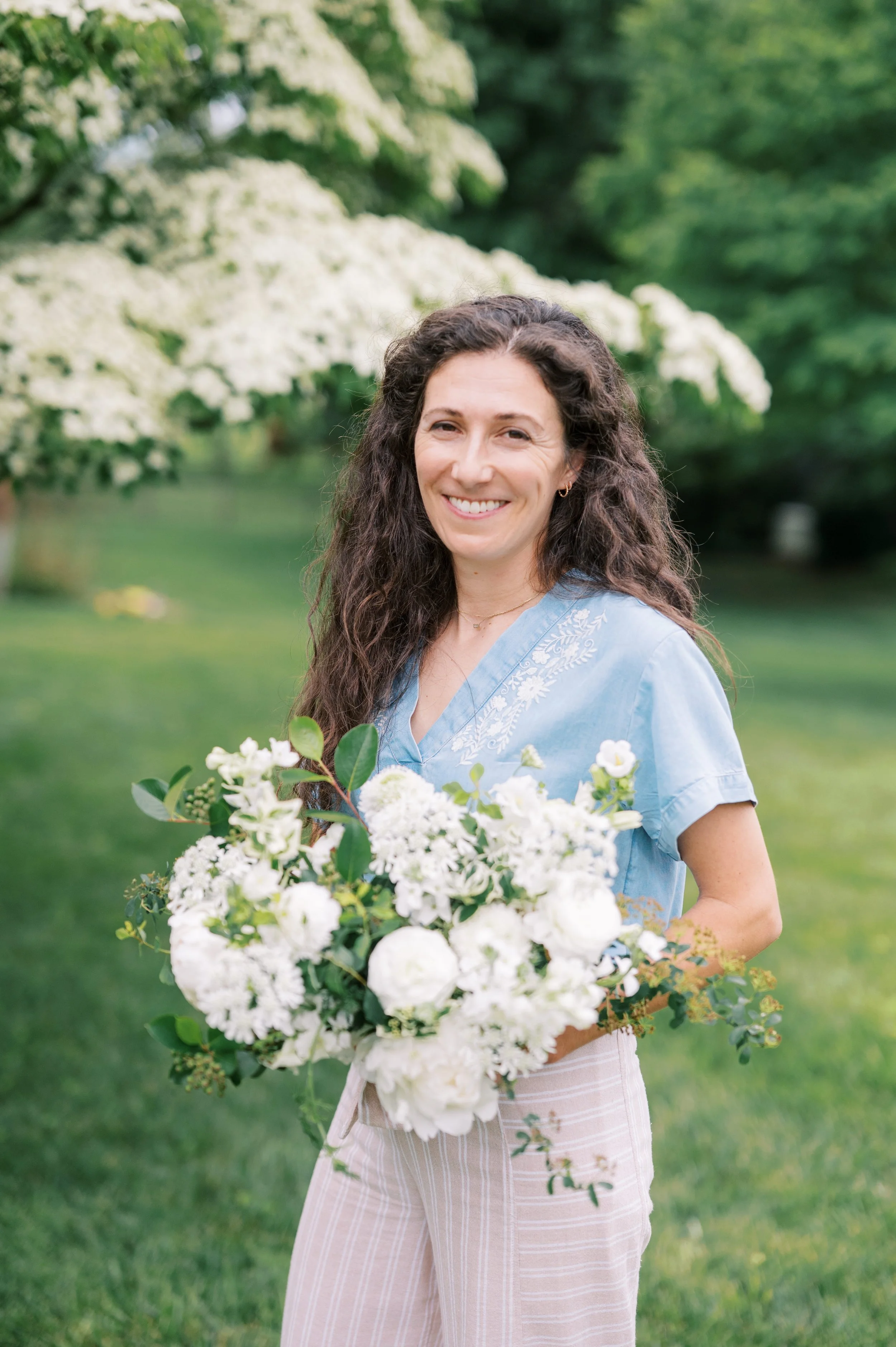 A woman with long, curly brown hair smiling outdoors in a park-like setting, holding a large bouquet of white flowers and greenery, wearing a light blue blouse with embroidery and beige striped pants.